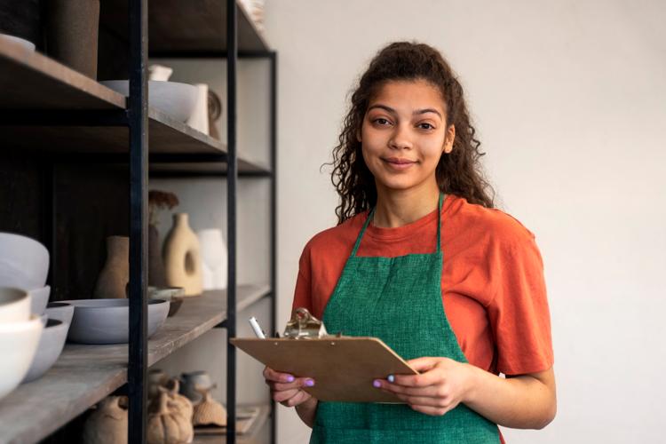Smiling woman wearing an apron holding a clipboard and pen standing in front of shelves full of pottery.